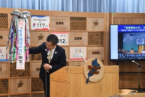 知事定例会見にて壇上で話す三日月知事の写真。知事の横に資料表示用の大きなモニターが映っている。