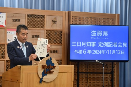 知事定例会見にて壇上で話す三日月知事の写真。知事の横に資料表示用の大きなモニターが映っている。