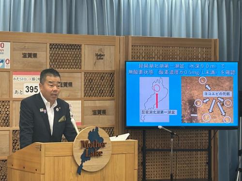 知事定例会見にて壇上で話す三日月知事の写真。知事の横に資料表示用の大きなモニターが映っている。