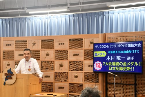 知事定例会見にて壇上で話す三日月知事の写真。知事の横に資料表示用の大きなモニターが映っている。
