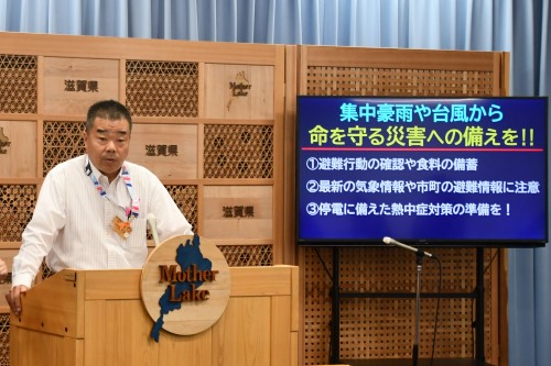 知事定例会見にて壇上で話す三日月知事の写真。知事の横に資料表示用の大きなモニターが映っている。