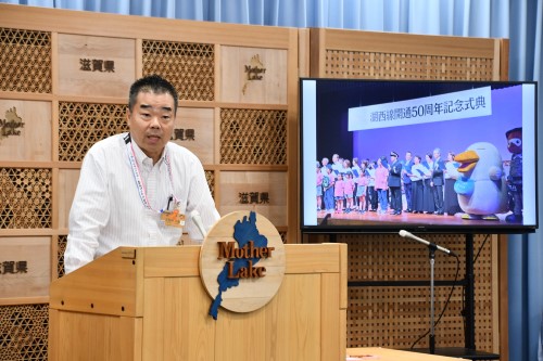 知事定例会見にて壇上で話す三日月知事の写真。知事の横に資料表示用の大きなモニターが映っている。