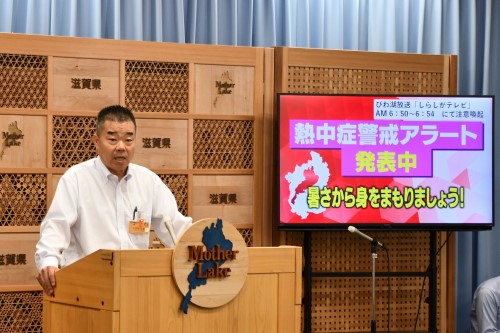 知事定例会見にて壇上で話す三日月知事の写真。知事の横に資料表示用の大きなモニターが映っている。
