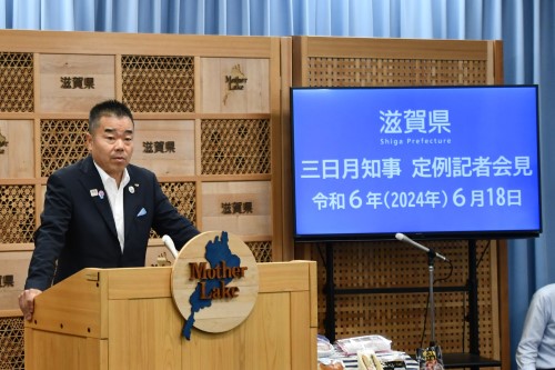 知事定例会見にて壇上で話す三日月知事の写真。知事の横に資料表示用の大きなモニターが映っている。