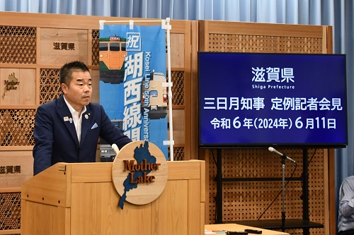 知事定例会見にて壇上で話す三日月知事の写真。知事の横に資料表示用の大きなモニターが映っている。