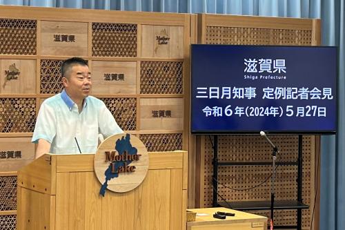 知事定例会見にて壇上で話す三日月知事の写真。知事の横に資料表示用の大きなモニターが映っている。