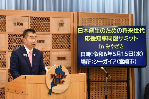 知事定例会見にて壇上で話す三日月知事の写真。日本創生のための将来世代応援知事同盟サミットInみやざきの情報(日時:令和6年5月15日(水曜日)開催、場所:シーガイヤ(宮崎市))が表示されている。