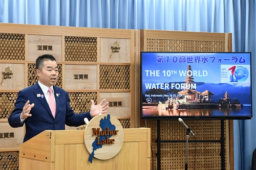 知事定例会見にて壇上で話す三日月知事の写真。話題提供の国際会議「世界水フォーラム」の情報がモニターに映っている。