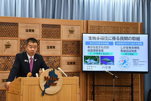 知事定例会見にて壇上で話題提供について話す三日月知事の写真。知事の横に大きなモニターが映っている。モニターには話題提供資料の一部が表示されている。